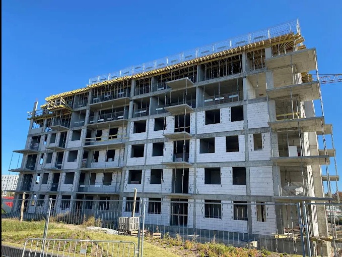 Wielopiętrowy budynek w stanie surowym otwartym, z widocznymi elementami konstrukcyjnymi i szalunkami na dachu, ogrodzony tymczasowym płotem.