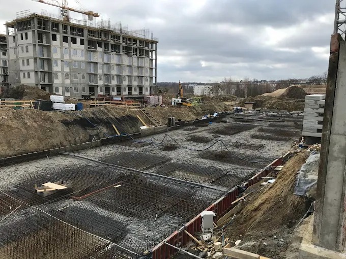 Zbrojenie fundamentów pod budynek wielorodzinny, widok z góry na siatkę zbrojeniową i szalunki. W tle budynek w stanie surowym i żuraw budowlany.