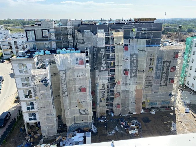 Wielopiętrowy budynek w trakcie budowy, osłonięty siatkami z logo producentów materiałów budowlanych, widoczne rusztowania i materiały budowlane na placu budowy.