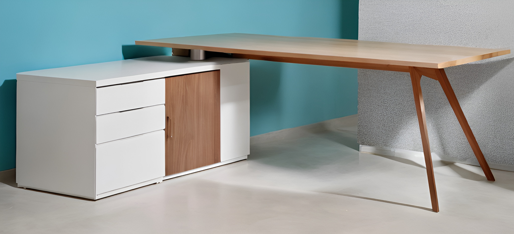 Nowoczesne biurko z jasnego drewna z trzema nogami, połączone z białą szafką z trzema szufladami i drewnianymi drzwiczkami, na tle turkusowej i szarej ściany.