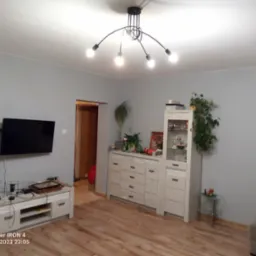 Jasne, szare ściany w pokoju z drewnianą podłogą, nowoczesna lampa sufitowa z pięcioma żarówkami, telewizor na ścianie nad białą szafką RTV, biała komoda z witryną i roślinami doniczkowymi.