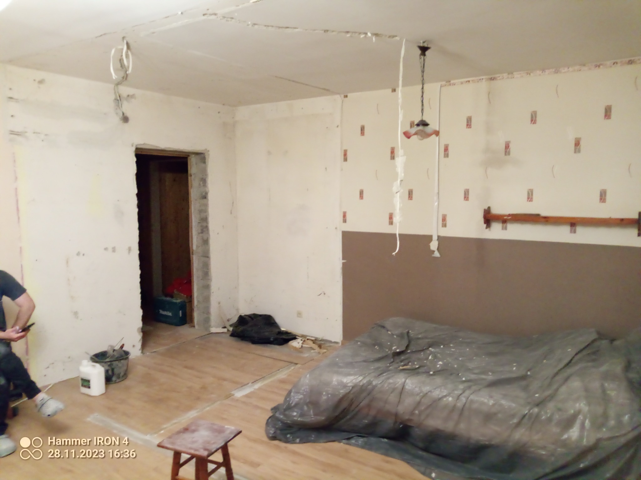 Wnętrze pokoju w Kisielicach w trakcie remontu: odsłonięte przewody elektryczne wiszące z sufitu, fragmenty starej tapety, podłoga częściowo przykryta folią ochronną, widoczne narzędzia i wiadro...
