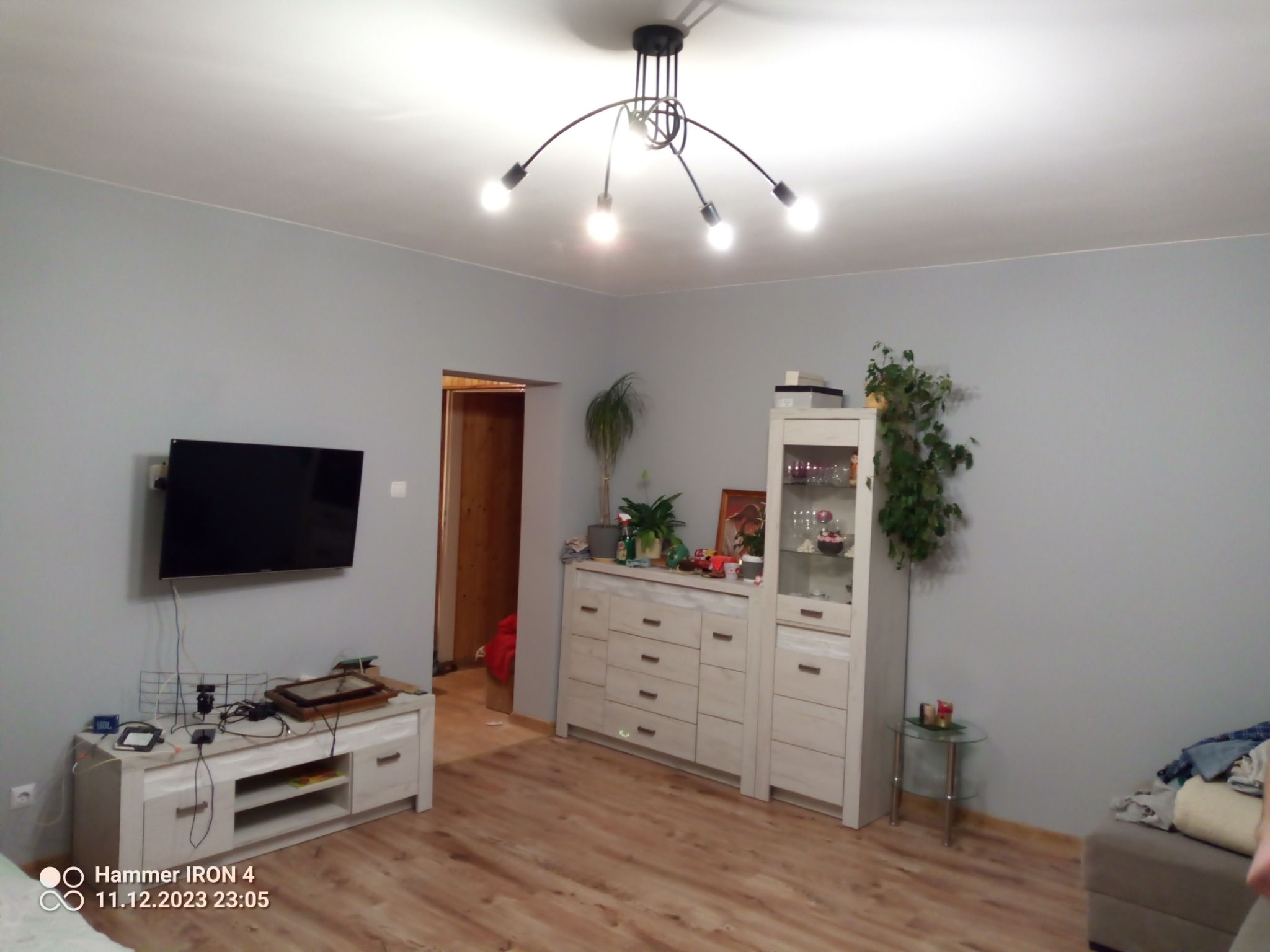 Jasne, szare ściany w pokoju z drewnianą podłogą, nowoczesna lampa sufitowa z pięcioma żarówkami, telewizor na ścianie nad białą szafką RTV, biała komoda z witryną i roślinami doniczkowymi.