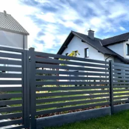 Nowoczesne, poziome przęsła palisadowe w kolorze grafitowym, zamontowane na metalowych słupkach z podmurówką, na tle domów jednorodzinnych i błękitnego nieba z chmurami. Widoczne logo producenta...