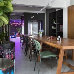 TENGO DESIGN - Nowoczesna aranżacja biura z długim drewnianym stołem, krzesłami w różnych stylach i neonowym oświetleniem. W tle widoczna strefa relaksu z nietypowym fotelem.
