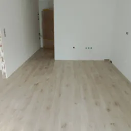 Jasne panele podłogowe ułożone w nowym, pustym pomieszczeniu z widocznymi gniazdkami elektrycznymi i otwartymi drzwiami w tle.