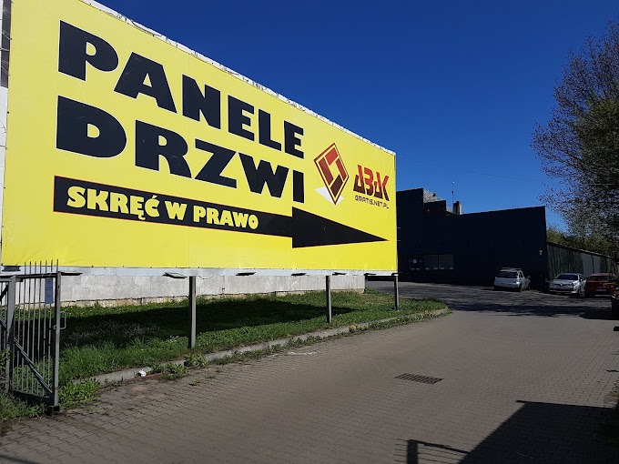 Żółty billboard reklamowy z napisem 'Panele Drzwi Skręć w Prawo' i logo firmy ABK Gratis.net.pl, ustawiony przy drodze dojazdowej do ciemnego budynku.
