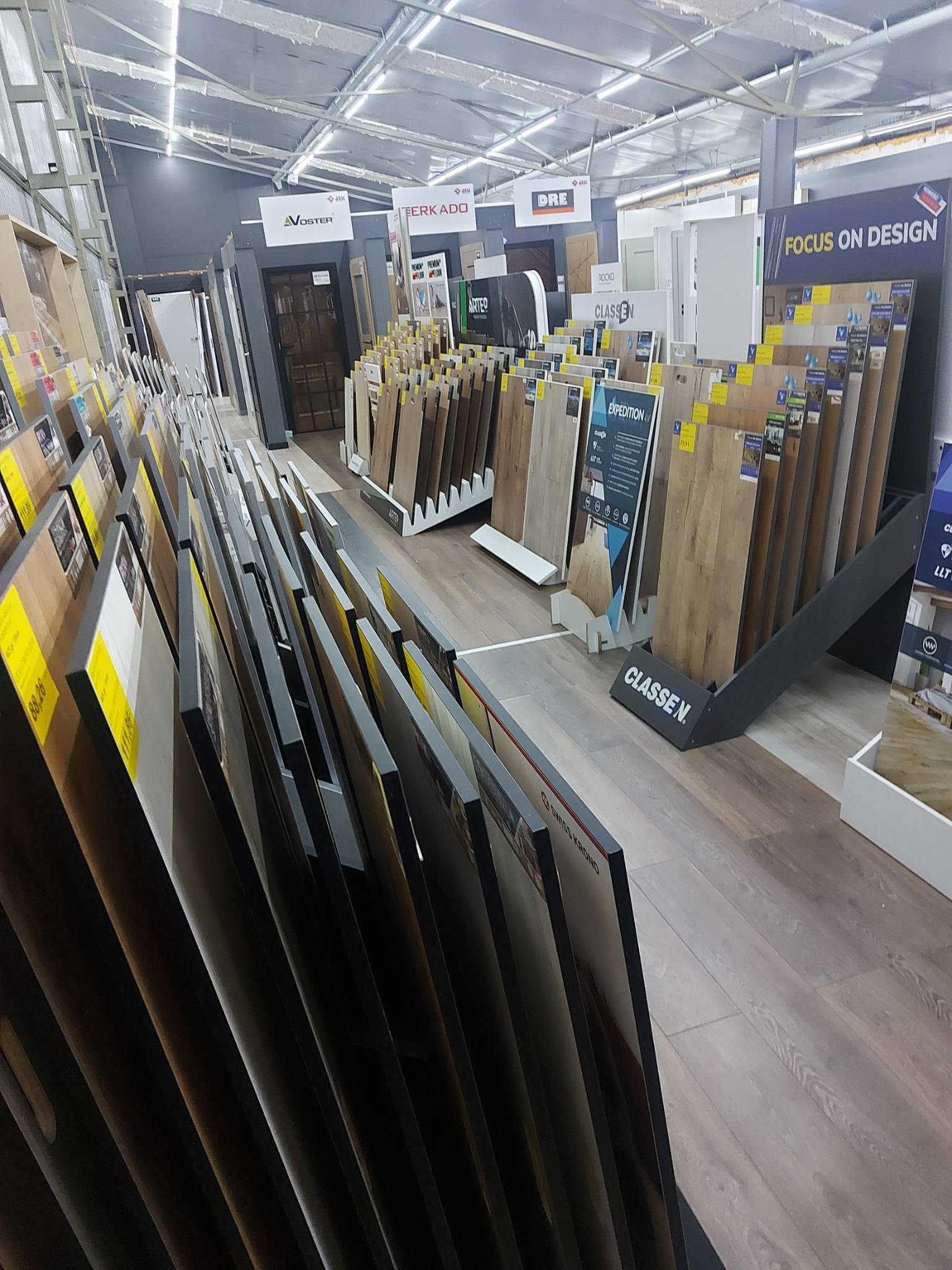 Ekspozycja różnorodnych paneli podłogowych i drzwi w sklepie, widoczne marki AVOSTER, TERKADO, DRE, CLASSEN, Swiss Krono, z cenami na ekspozytorach, oświetlenie sufitowe.