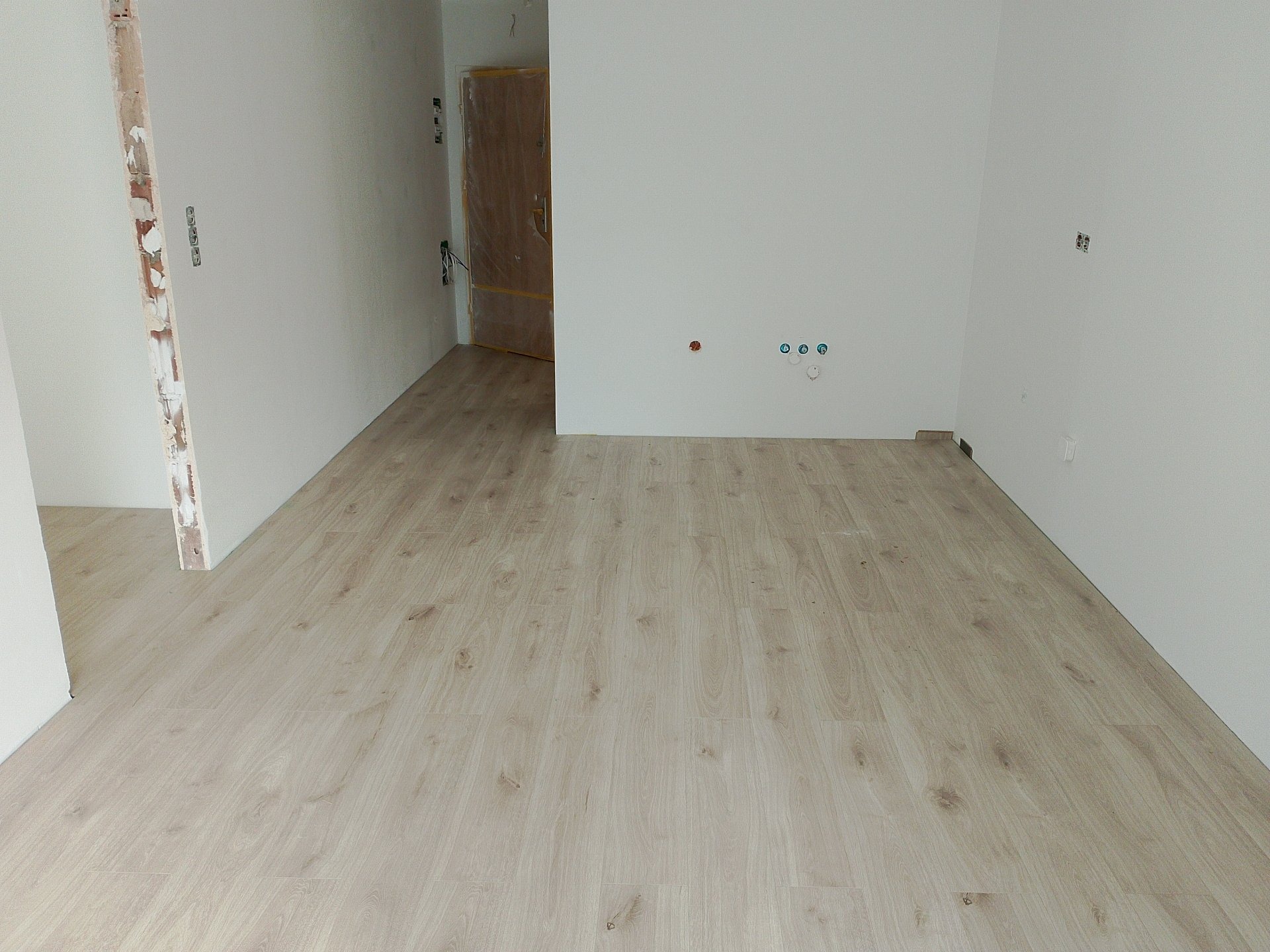 Jasne panele podłogowe ułożone w nowym, pustym pomieszczeniu z widocznymi gniazdkami elektrycznymi i otwartymi drzwiami w tle.