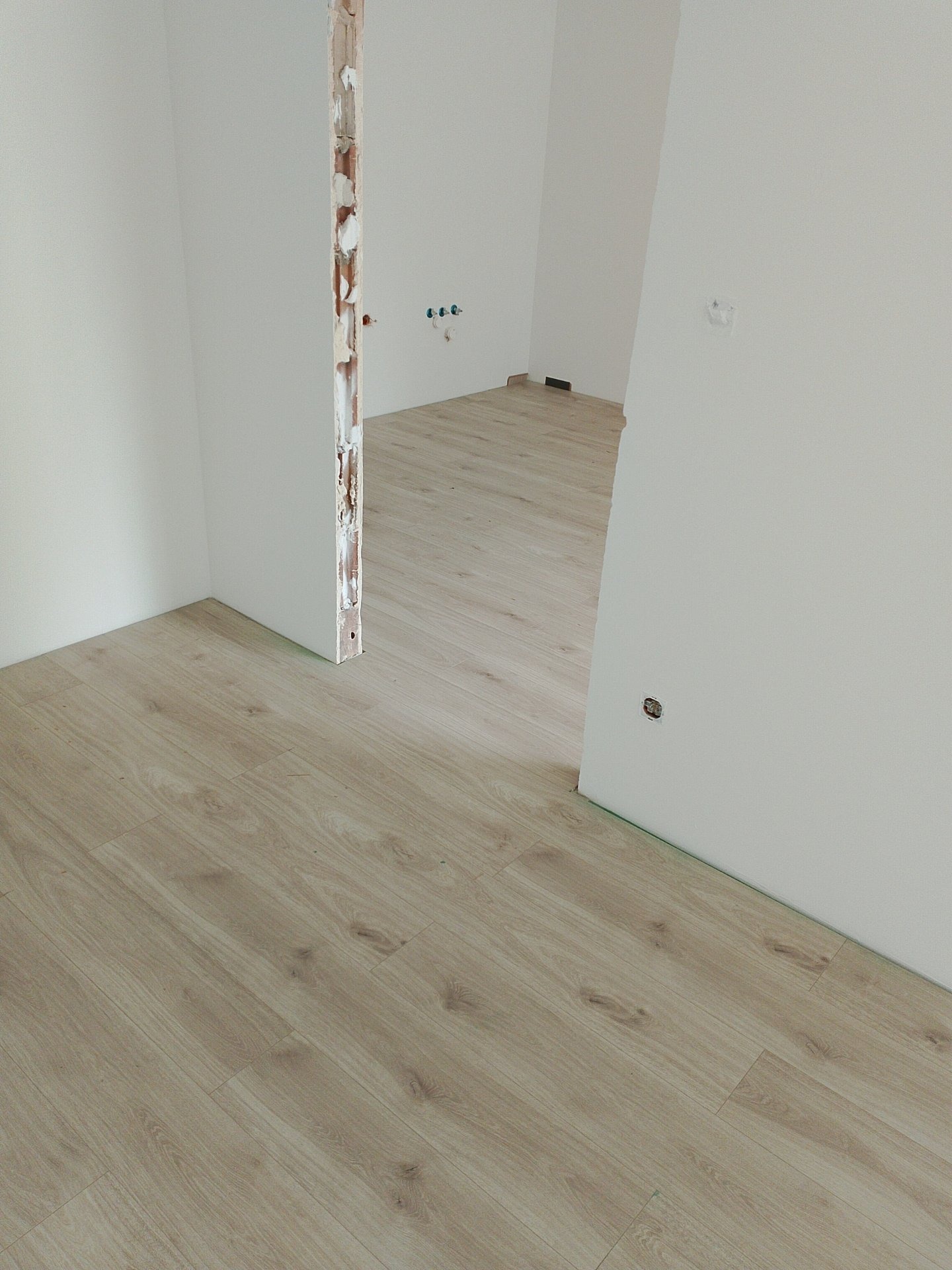 Jasne panele podłogowe ułożone w pustym, odmalowanym pokoju z widoczną surową ościeżnicą i przygotowanymi gniazdkami elektrycznymi.