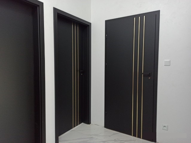 Nowoczesne, czarne drzwi wewnętrzne z trzema złotymi paskami dekoracyjnymi, widoczne w korytarzu z marmurową podłogą.