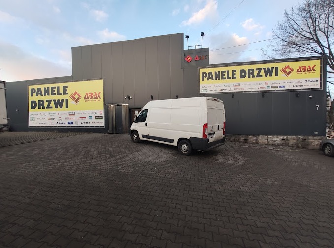 Zewnętrzny widok budynku firmy z szarymi ścianami, reklamującej panele i drzwi, z logo firmy i białym vanem zaparkowanym przed wejściem.