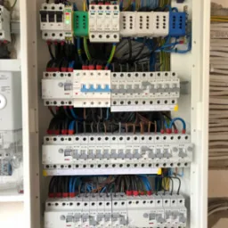 Rozdzielnica elektryczna w trakcie montażu, widoczne bezpieczniki, przewody w różnych kolorach, listwy zaciskowe i elementy montażowe, ściana w tle z widocznymi śladami prac wykończeniowych.