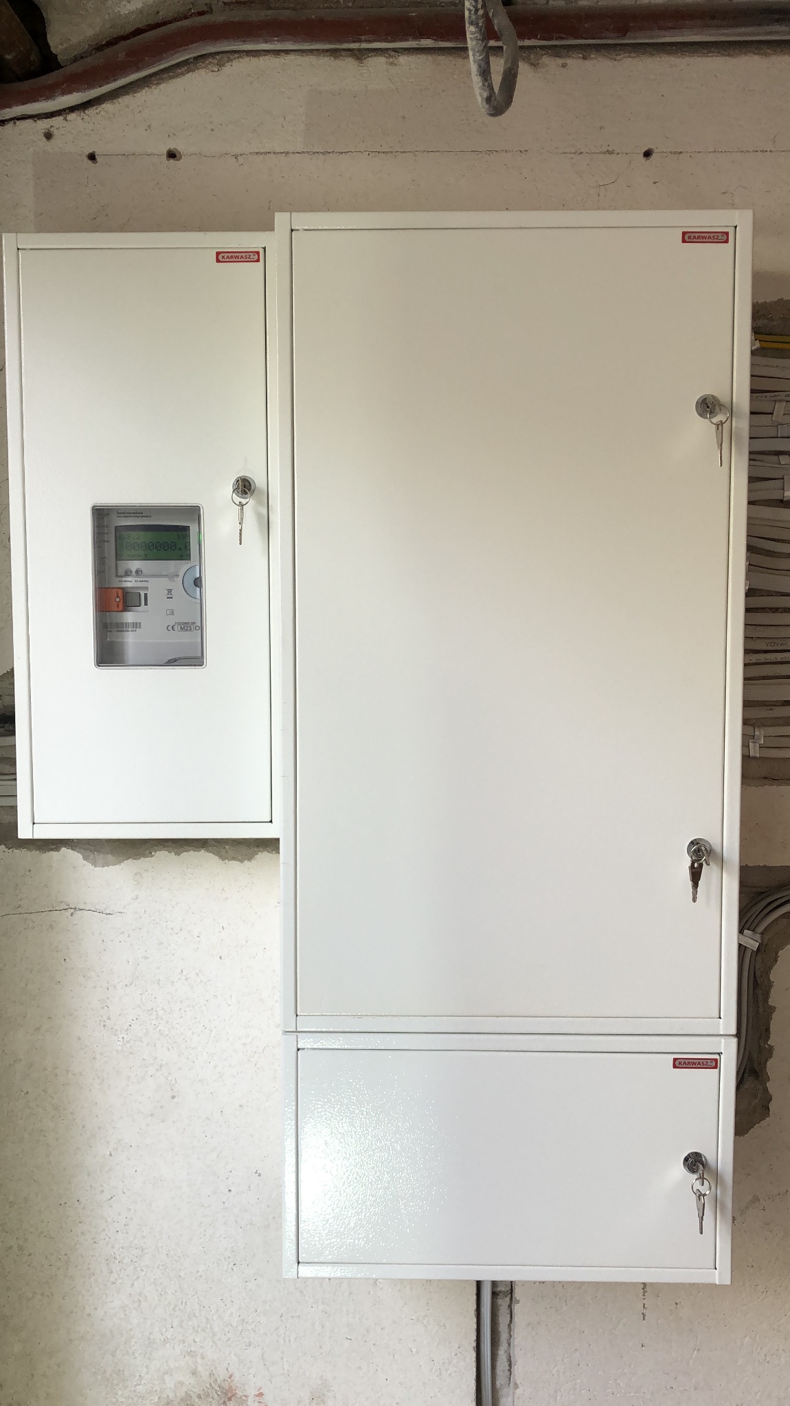 Biała, metalowa szafka elektryczna z licznikiem i zamkami, zamontowana na surowej, betonowej ścianie w trakcie remontu. Widoczne kable i rury pod sufitem.