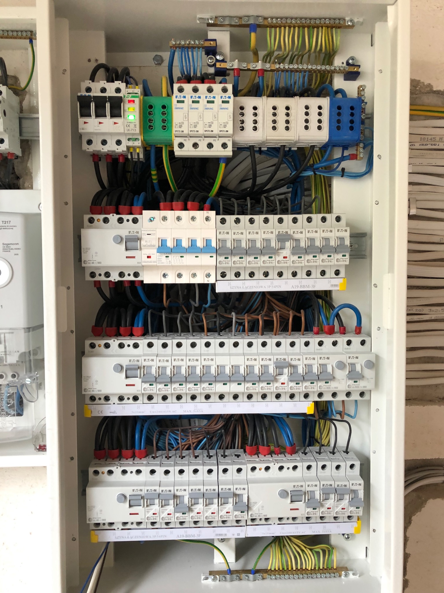 Rozdzielnica elektryczna w trakcie montażu, widoczne bezpieczniki, przewody w różnych kolorach, listwy zaciskowe i elementy montażowe, ściana w tle z widocznymi śladami prac wykończeniowych.