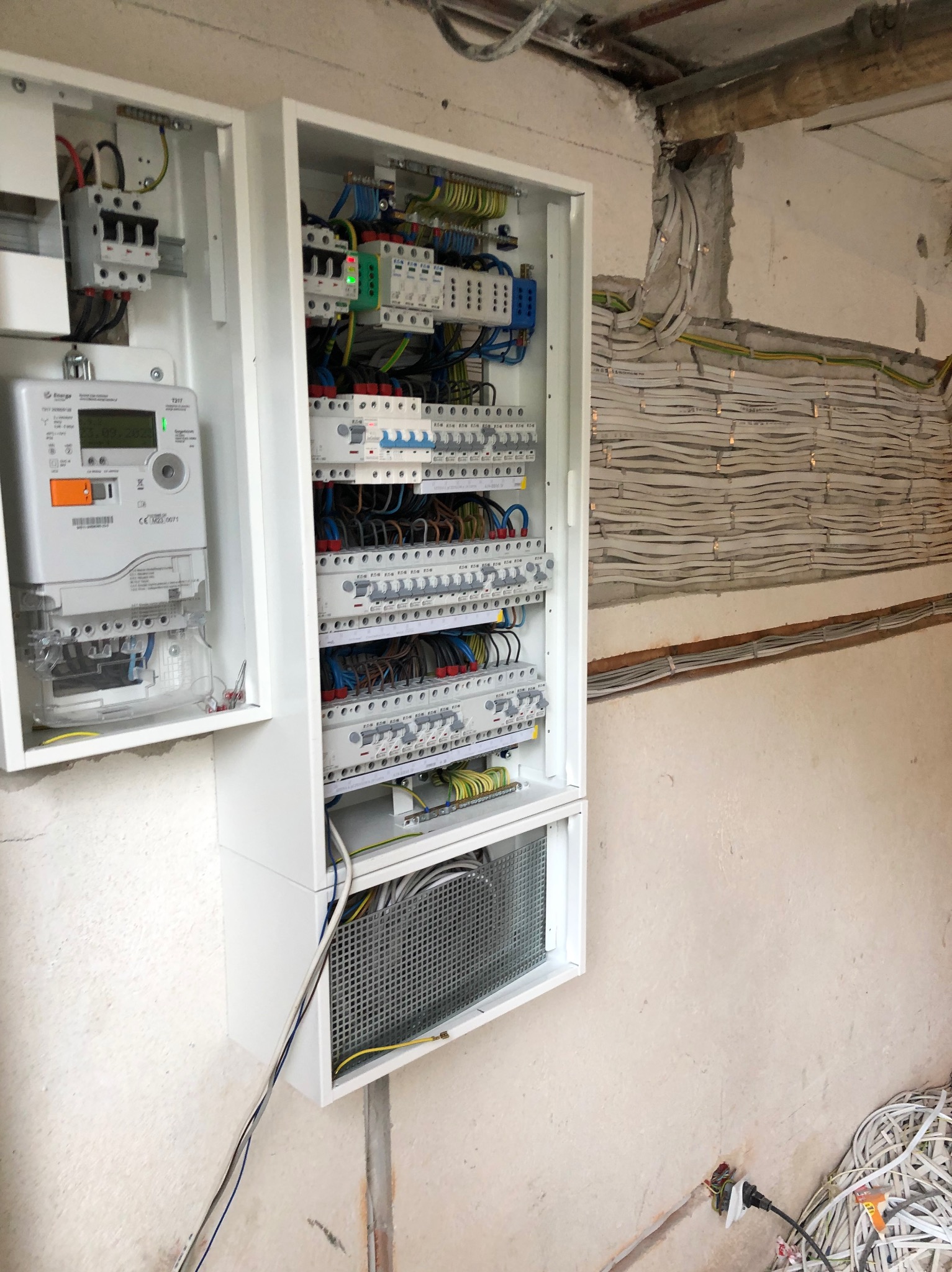 Otwarta biała skrzynka rozdzielcza z licznikiem i zabezpieczeniami, widoczne kable w ścianie i na podłodze w surowym stanie.