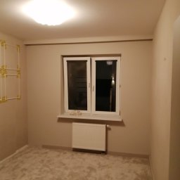 Vielkibud Oleksandr Kulbakov - Pokój po odświeżeniu: beżowe ściany, białe okno z parapetem, grzejnik pod oknem, jasny dywan na podłodze, dekoracyjne elementy na ścianie po lewej.