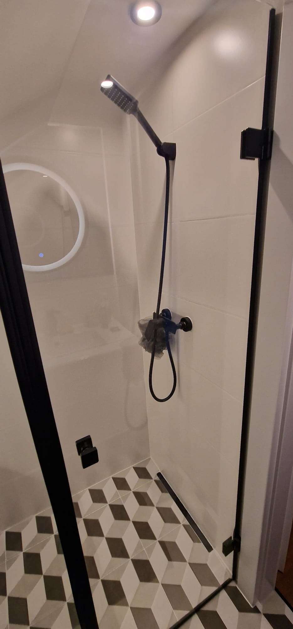 Nowoczesna kabina prysznicowa z czarną armaturą, geometryczną posadzką i okrągłym lustrem z podświetleniem LED.