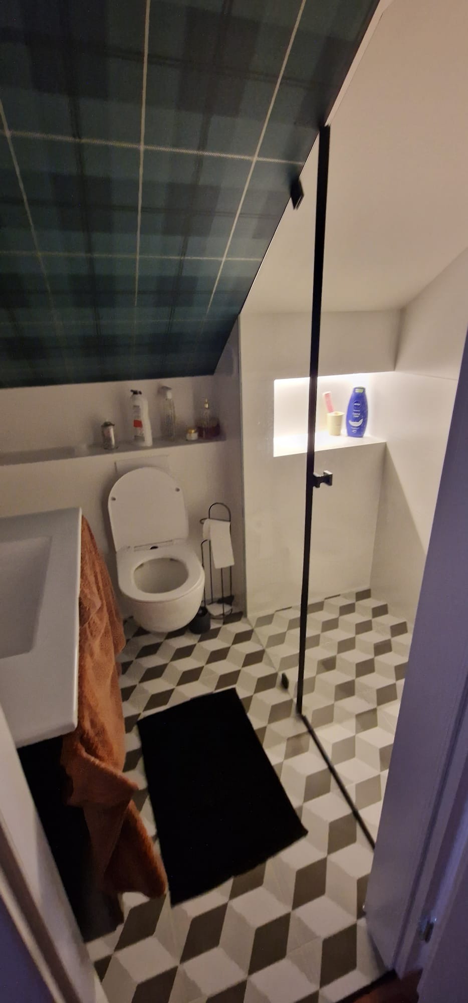 Ukończona łazienka w poddaszu z wzorzystą tapetą w kratę na suficie, geometrycznymi płytkami podłogowymi, kabiną prysznicową typu walk-in z czarnymi profilami, białą umywalką i toaletą oraz wnęką...