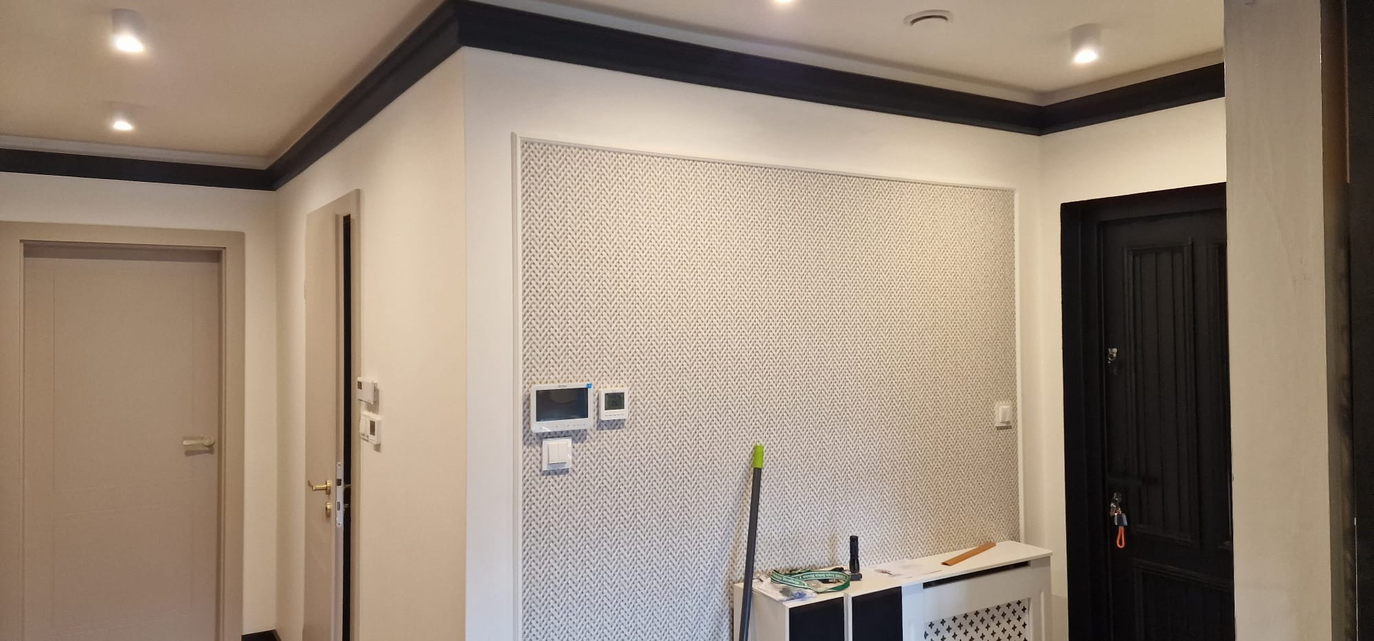 Wykończone wnętrze przedpokoju z dekoracyjną tapetą w geometryczny wzór, ciemną listwą przysufitową i czarnymi drzwiami wejściowymi, widoczne akcesoria malarskie.