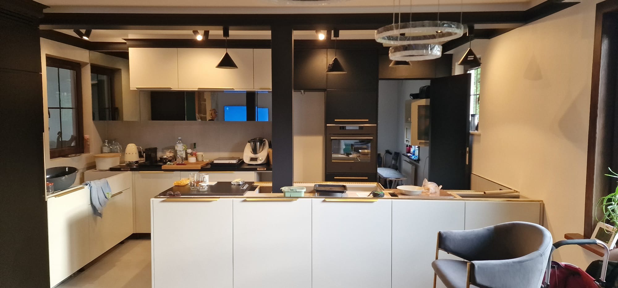 Nowoczesna kuchnia z wyspą, fronty szafek w kolorze białym i czarnym, złote uchwyty, oświetlenie punktowe i wiszące lampy ring, widoczne akcesoria kuchenne i sprzęt AGD.