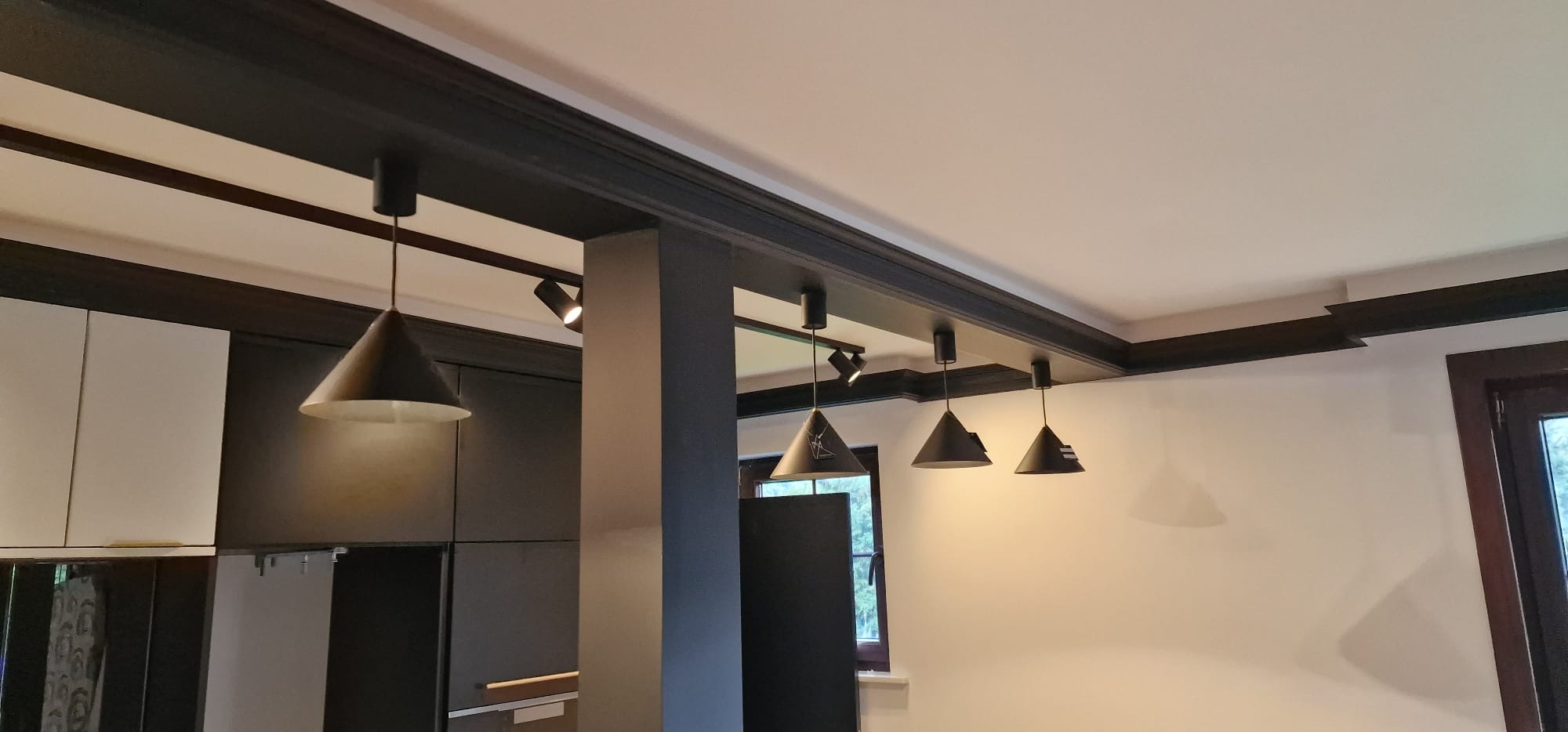 Nowoczesne oświetlenie w kuchni: czarne, stożkowe lampy wiszące pod ciemnym, drewnianym sufitem podwieszanym z dekoracyjnymi listwami, widoczna zabudowa kuchenna w odcieniach bieli i czerni.