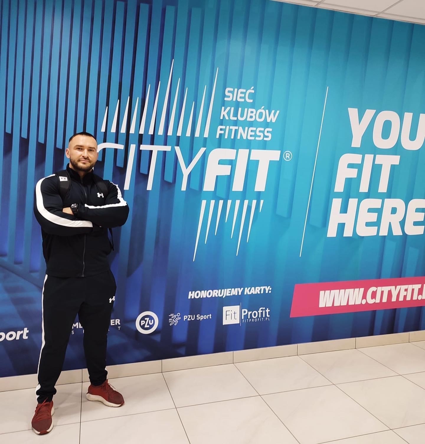 Mężczyzna w sportowym stroju stoi na tle ściany z logo sieci klubów fitness CityFit, prezentując ofertę honorowania kart PZU Sport i Fit Profit.