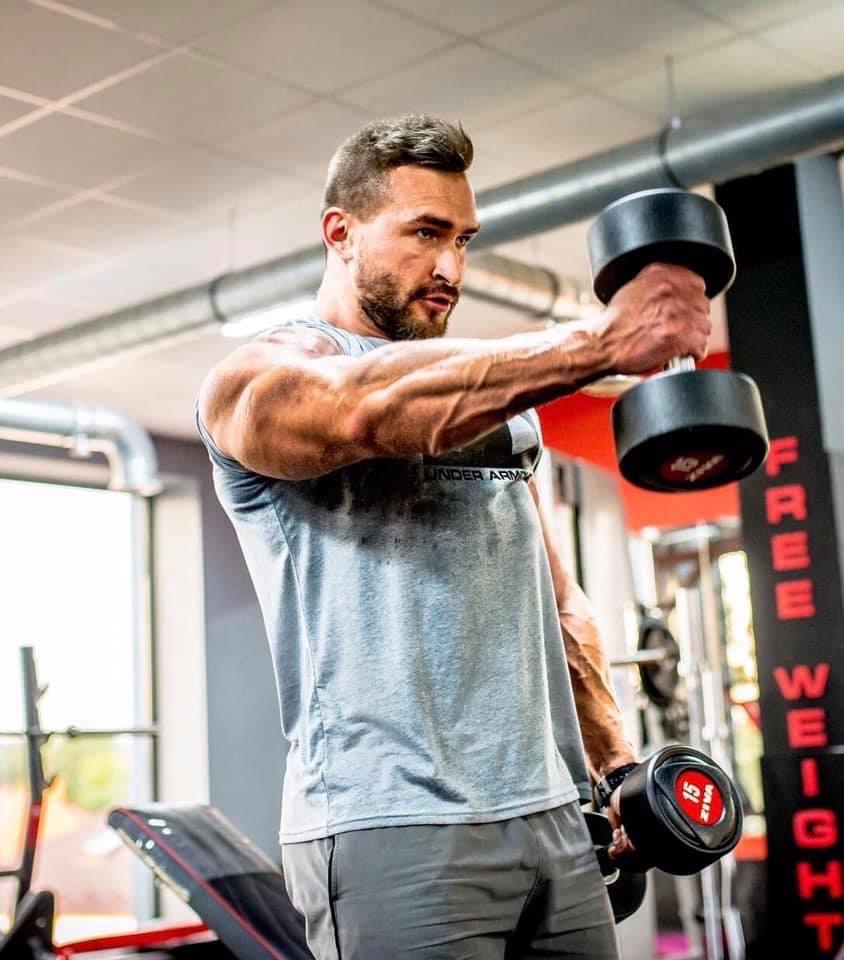 Mężczyzna z brodą w szarej koszulce Under Armour ćwiczy unoszenie hantli w przód na siłowni, trzymając w jednej ręce hantlę 15 kg, a w drugiej 10 kg.