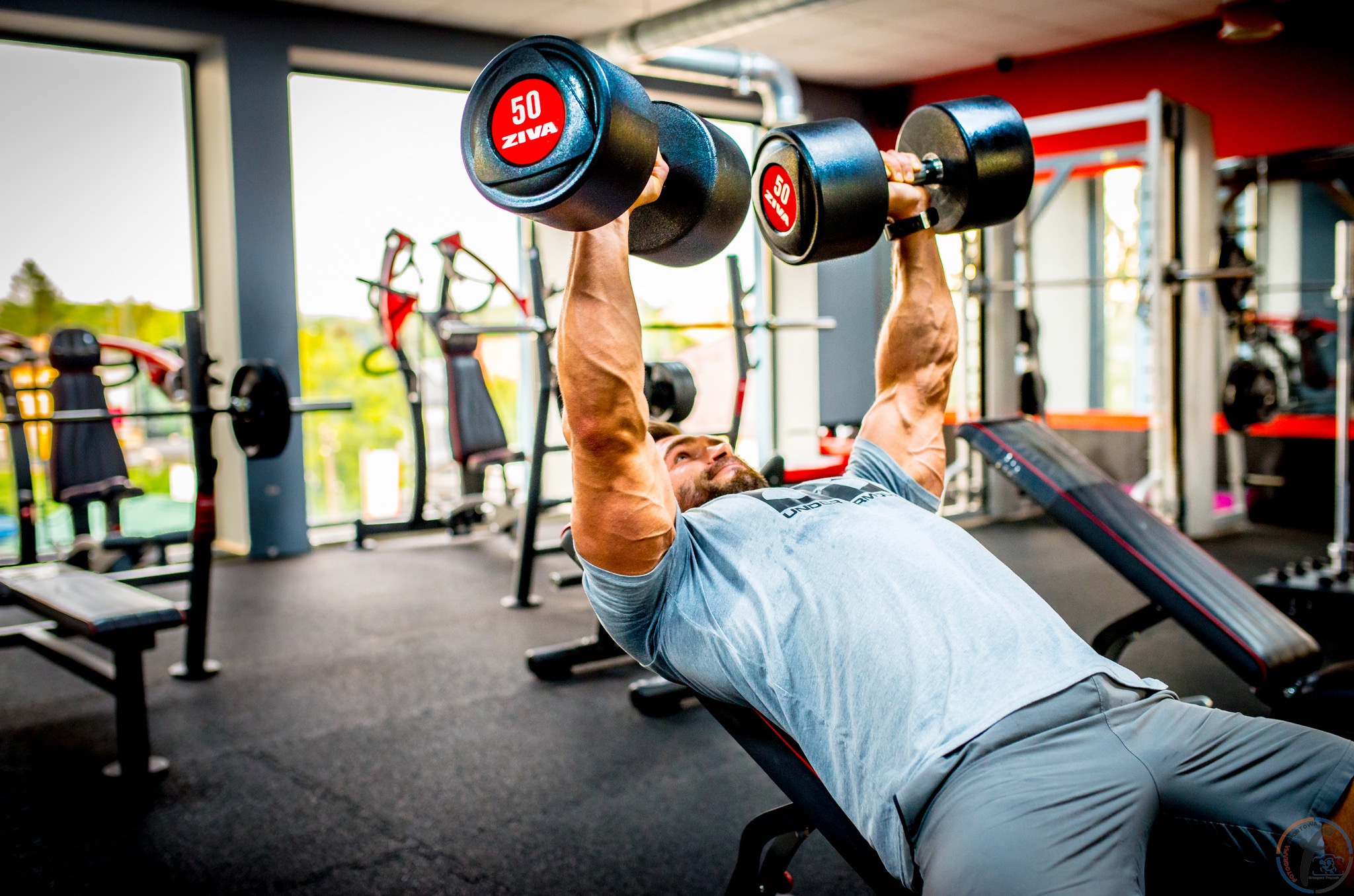 Mężczyzna leżący na ławce w siłowni, podnosi ciężkie hantle ZIVA o wadze 50 kg, z widocznymi żyłami na ramionach, w tle sprzęt do ćwiczeń i okno.