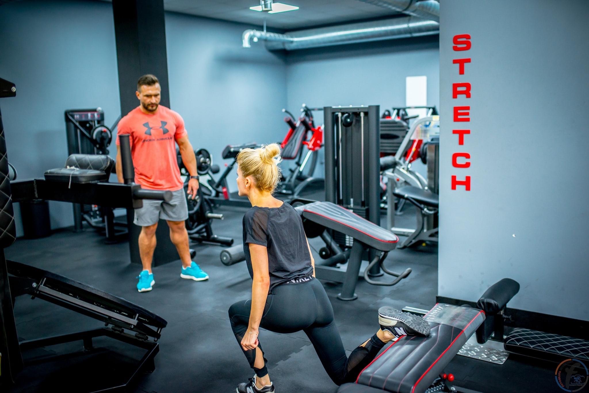 Kobieta w czarnym stroju ćwiczy rozciąganie w siłowni, opierając nogę o ławkę, a w tle stoi mężczyzna w sportowym ubraniu i obserwuje.
