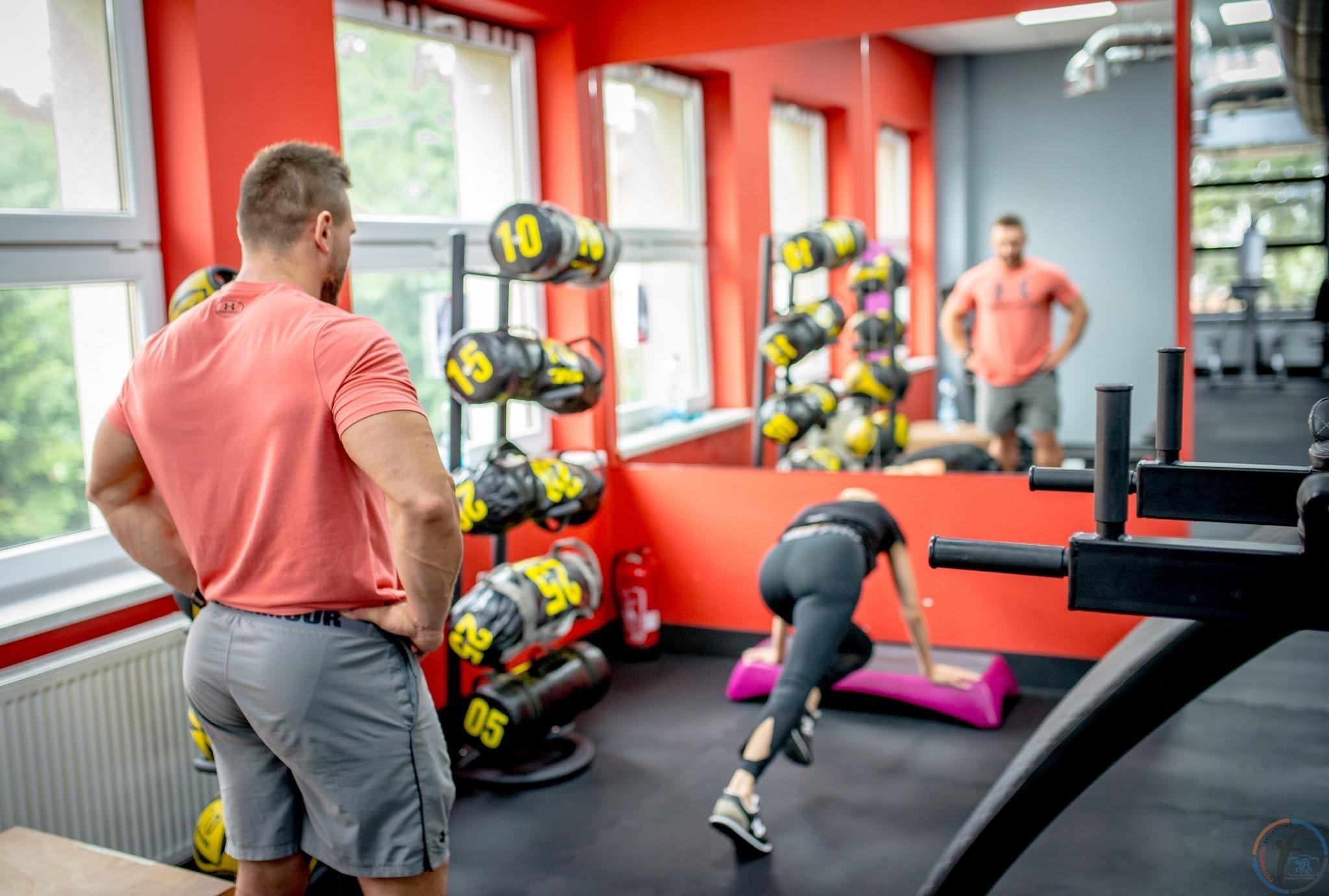 Mężczyzna w pomarańczowej koszulce obserwuje ćwiczącą kobietę na stepie w siłowni z czerwonymi ścianami, widoczne worki treningowe z żółtymi numerami.