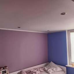 USŁUGI BUDOWLANO-REMONTOWE DAWID NAKIELSKI - Pokój dziecięcy po malowaniu ścian na fioletowo i niebiesko. Widoczne oświetlenie sufitowe, okno z roletą i dziecięce dekoracje na półce. Ściany pomalowane na dwa kolory.