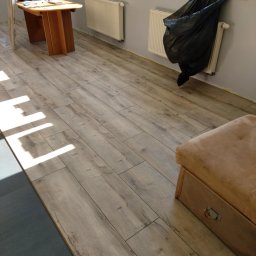 USŁUGI BUDOWLANO-REMONTOWE DAWID NAKIELSKI - Ułożone panele podłogowe w jasnym odcieniu drewna, widoczne narzędzia pracy i fragment mebla. Częściowo widoczny podkład pod panele.