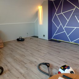 USŁUGI BUDOWLANO-REMONTOWE DAWID NAKIELSKI - Pokój na poddaszu po remoncie: świeżo ułożone panele podłogowe, pomalowana ściana w geometryczne wzory. Widoczny sprzęt do sprzątania i wózek transportowy.