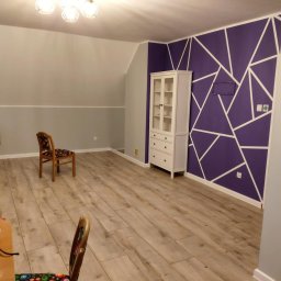 USŁUGI BUDOWLANO-REMONTOWE DAWID NAKIELSKI - Pokój po odświeżeniu: fioletowa ściana z białymi geometrycznymi wzorami, jasna podłoga, biała komoda. Widoczne krzesła i fragment stołu z butelką.