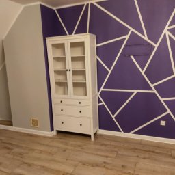 USŁUGI BUDOWLANO-REMONTOWE DAWID NAKIELSKI - Pokój po odświeżeniu: fioletowa ściana z białymi geometrycznymi wzorami, biała komoda, panele podłogowe. Widoczne gniazdka elektryczne i włączniki.