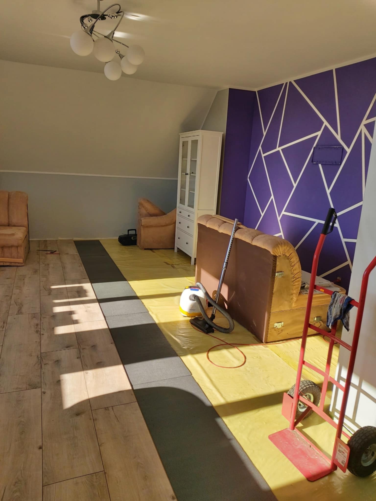 Fragment pokoju w trakcie prac wykończeniowych: ułożona podłoga z paneli, zabezpieczona folią, ściana z geometrycznym wzorem. Widoczny odkurzacz i wózek transportowy.