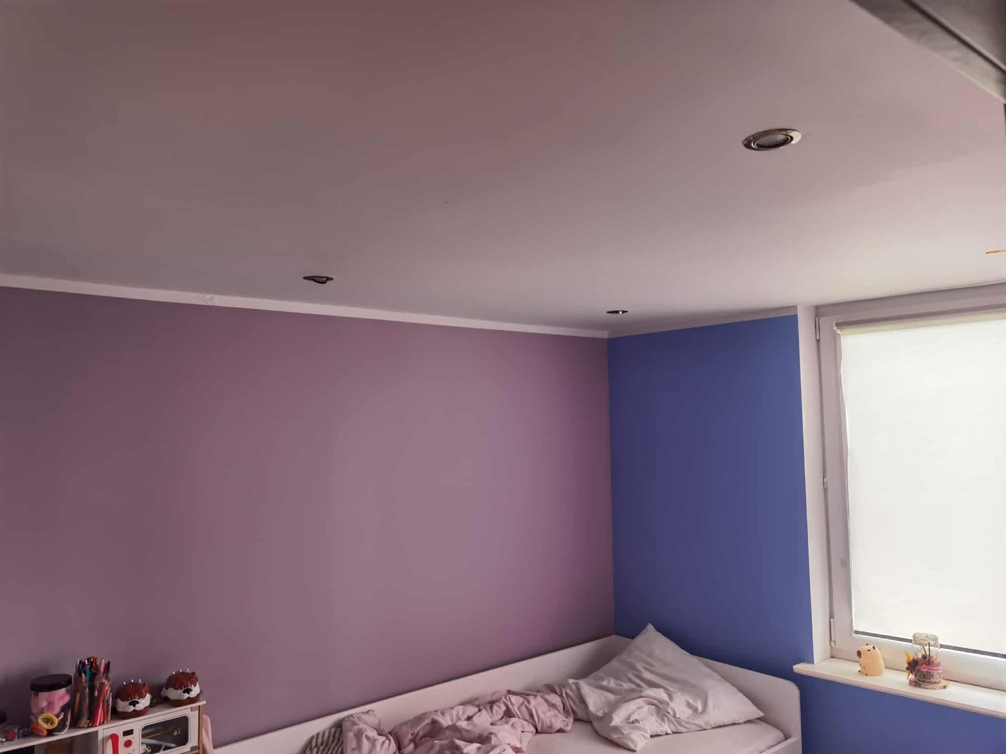 Pokój dziecięcy po malowaniu ścian na fioletowo i niebiesko. Widoczne oświetlenie sufitowe, okno z roletą i dziecięce dekoracje na półce. Ściany pomalowane na dwa kolory.