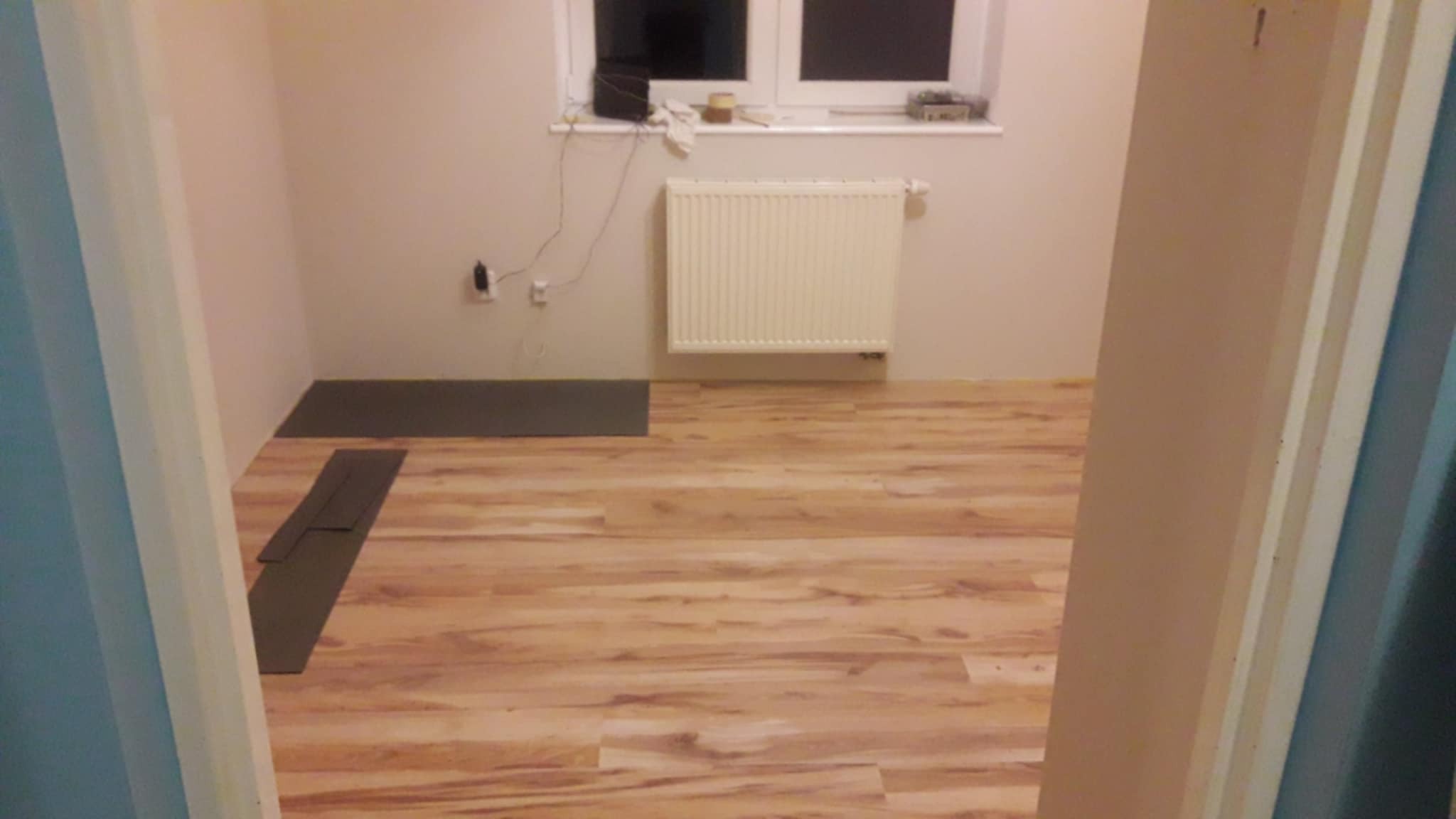 Fragment pokoju z ułożoną podłogą z paneli w odcieniach drewna. Widoczne panele podkładowe i grzejnik pod oknem. Ujęcie zza framugi drzwi.