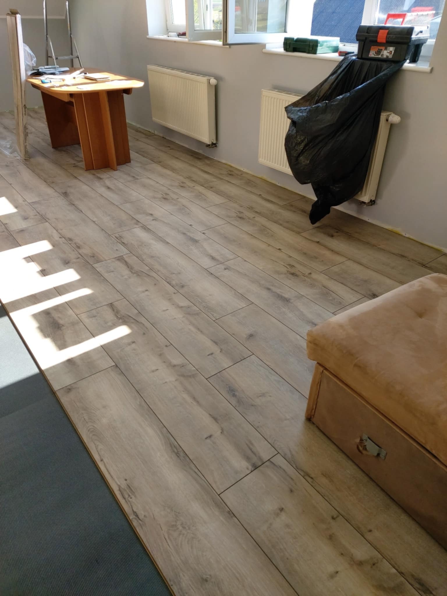 Ułożone panele podłogowe w jasnym odcieniu drewna, widoczne narzędzia pracy i fragment mebla. Częściowo widoczny podkład pod panele.