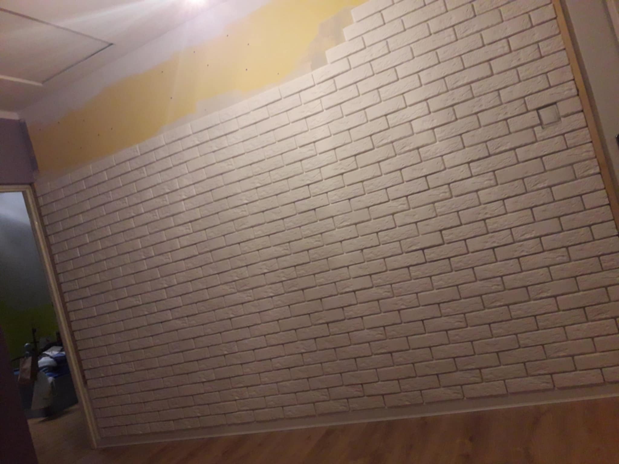 Ściana wyłożona beżową cegłą dekoracyjną w trakcie prac wykończeniowych. Widoczny fragment pomalowanej ściany i drewniana podłoga. Ujęcie z dołu.