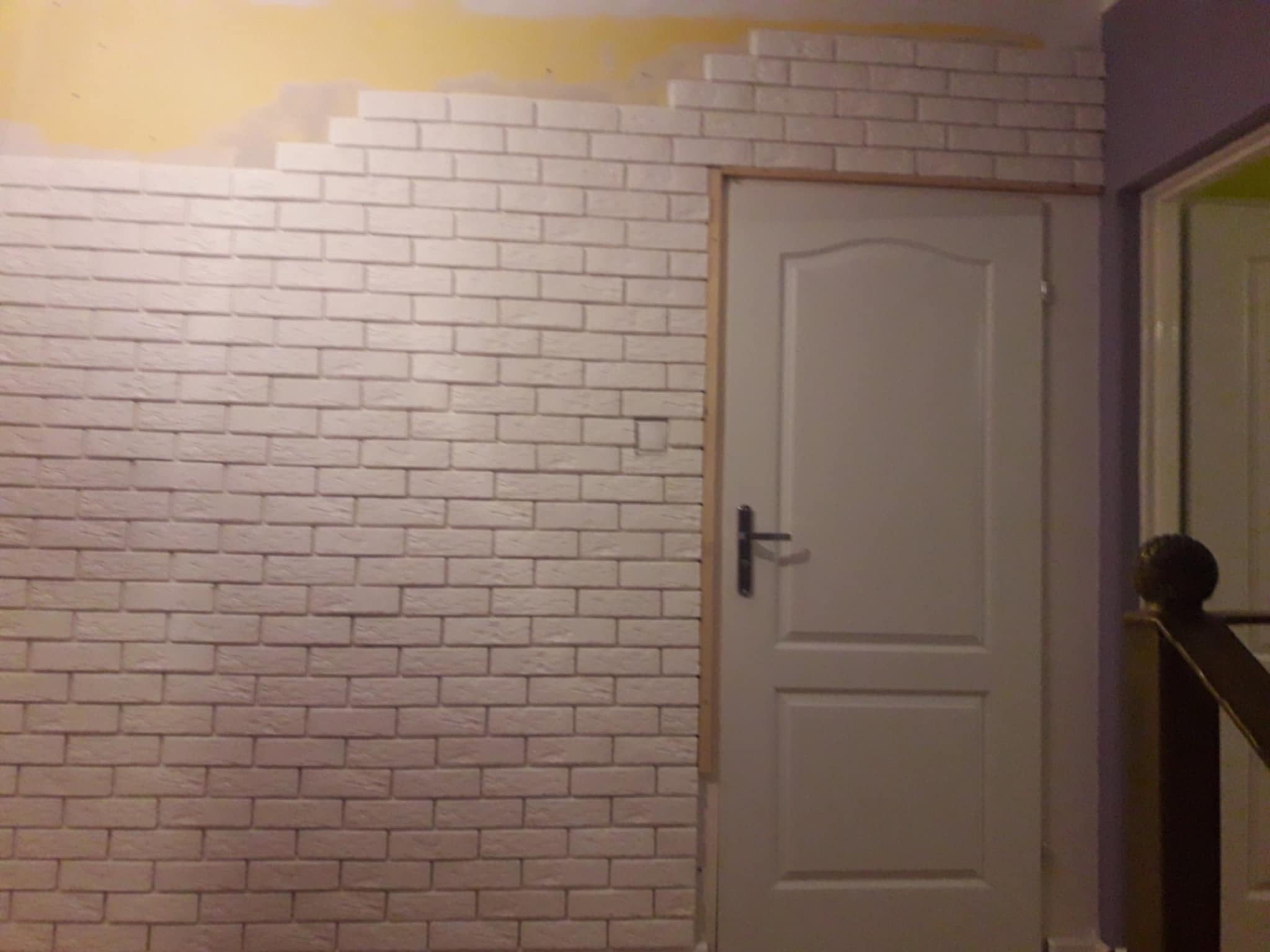 Ściana z białej cegły dekoracyjnej w trakcie montażu, widoczne niedokończone fragmenty i rama drzwi. Górna część ściany w kolorze żółtym, kontrastująca z bielą cegieł.