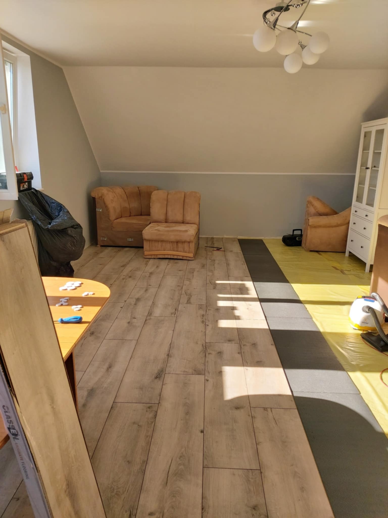 Montaż paneli podłogowych w pokoju na poddaszu. Częściowo ułożone panele, widoczne narzędzia i materiały przygotowane do dalszej pracy. W tle meble i okno.