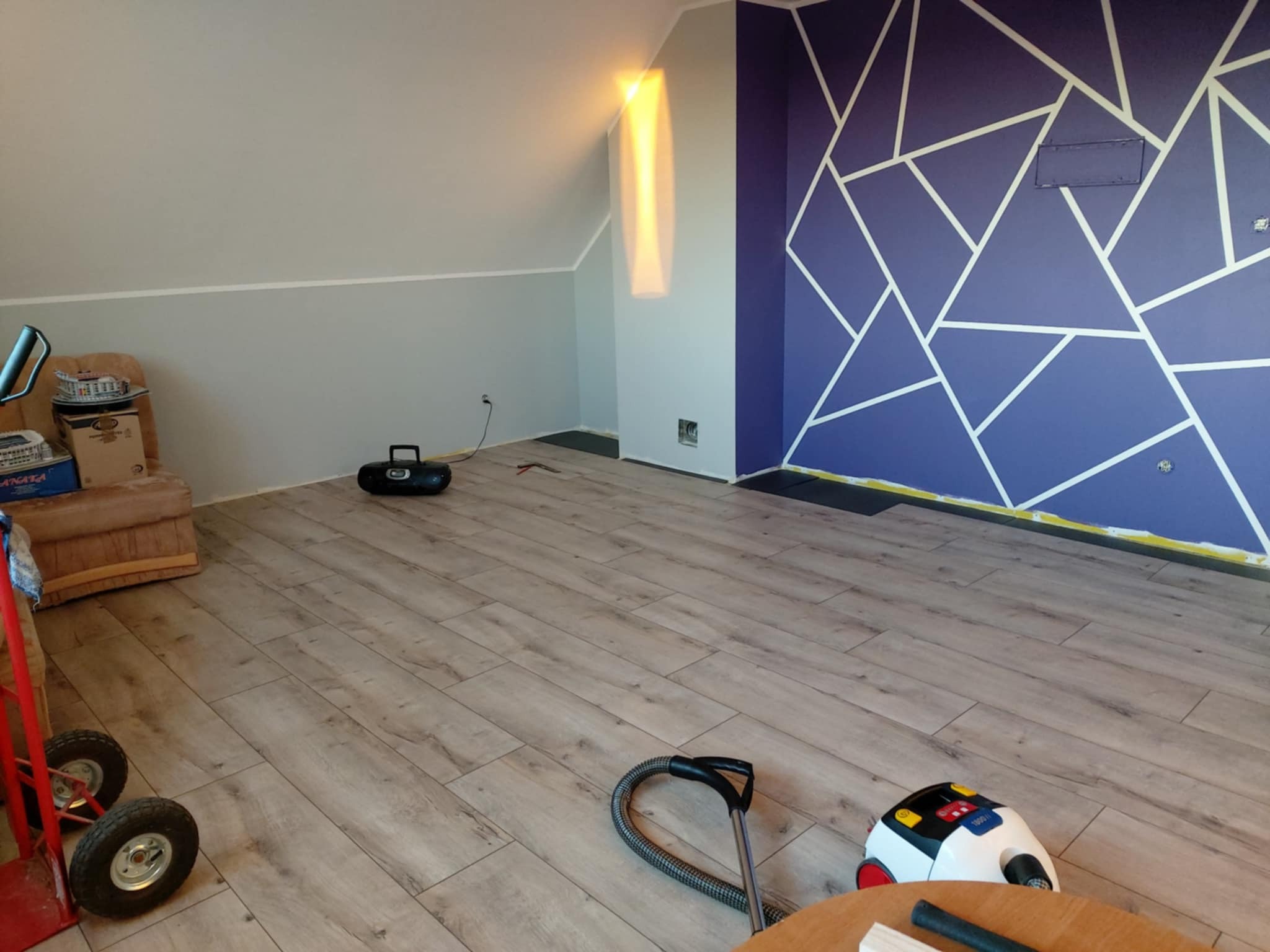 Pokój na poddaszu po remoncie: świeżo ułożone panele podłogowe, pomalowana ściana w geometryczne wzory. Widoczny sprzęt do sprzątania i wózek transportowy.