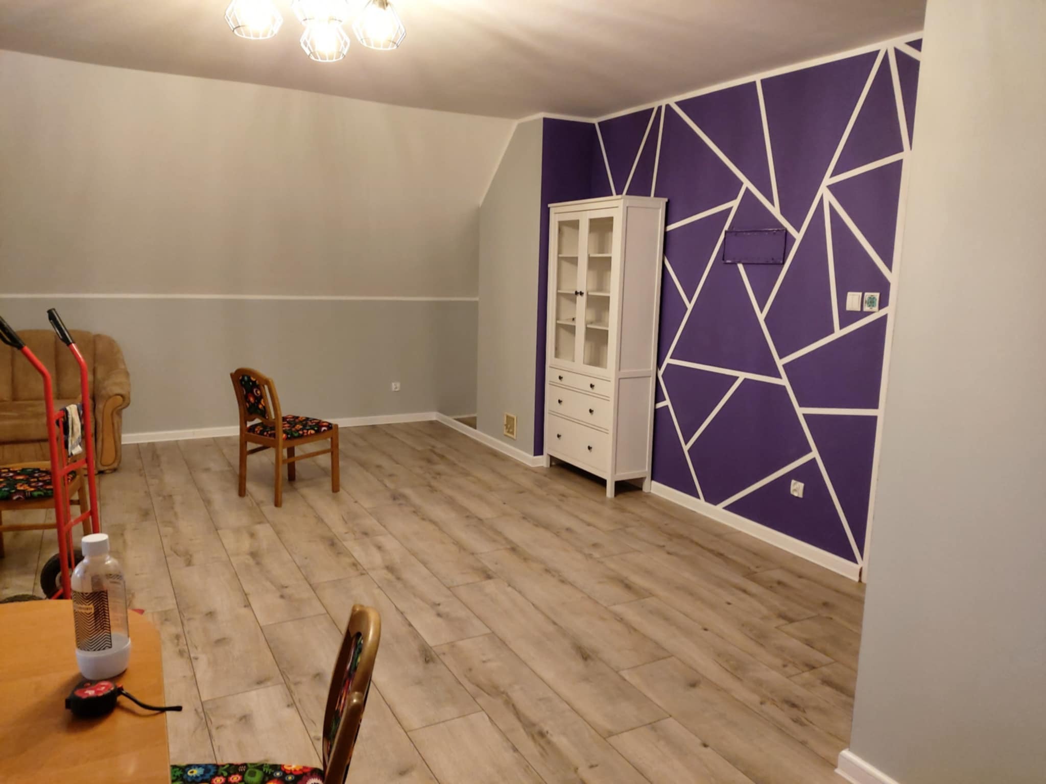 Pokój po odświeżeniu: fioletowa ściana z białymi geometrycznymi wzorami, jasna podłoga, biała komoda. Widoczne krzesła i fragment stołu z butelką.
