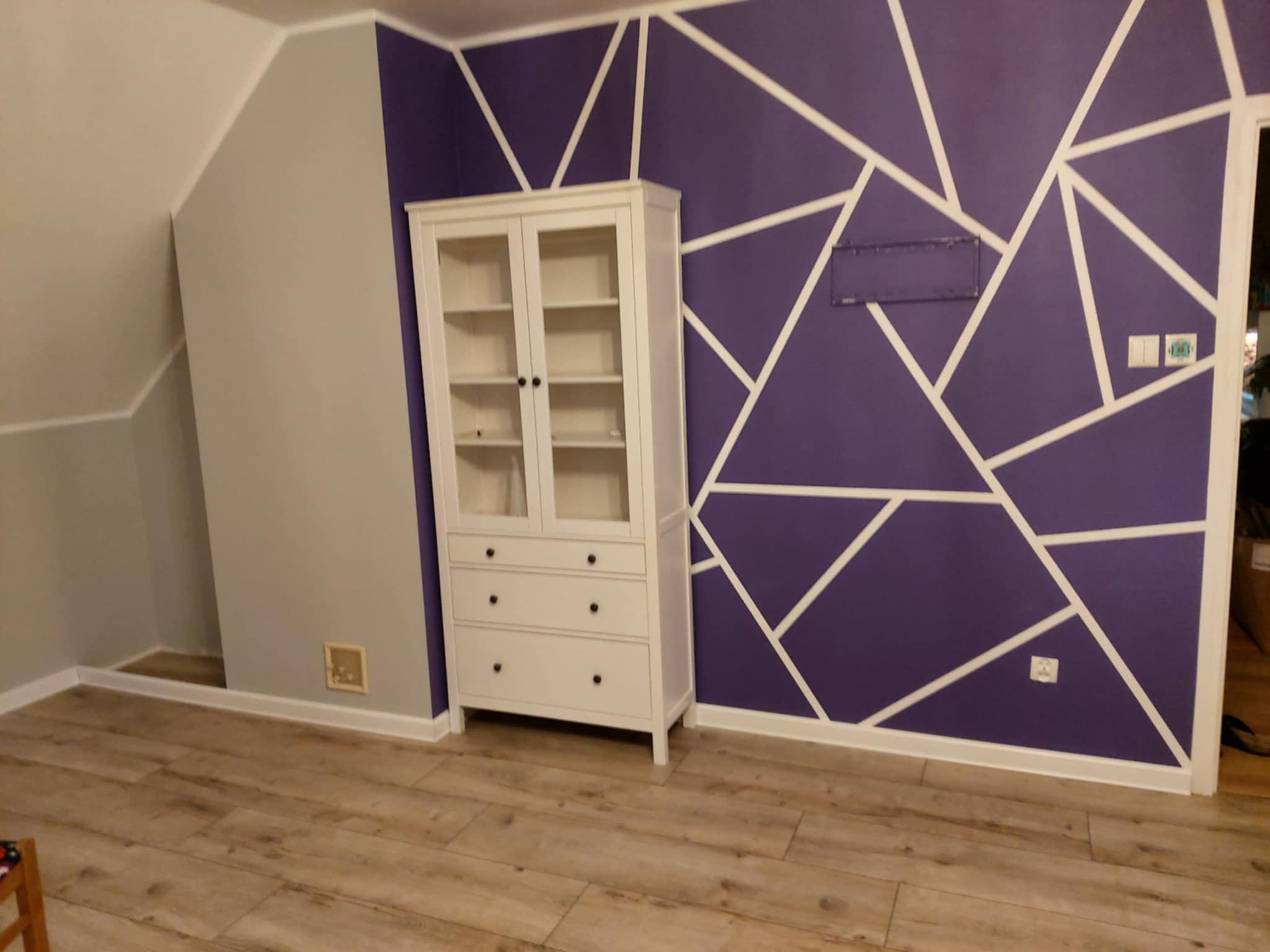 Pokój po odświeżeniu: fioletowa ściana z białymi geometrycznymi wzorami, biała komoda, panele podłogowe. Widoczne gniazdka elektryczne i włączniki.