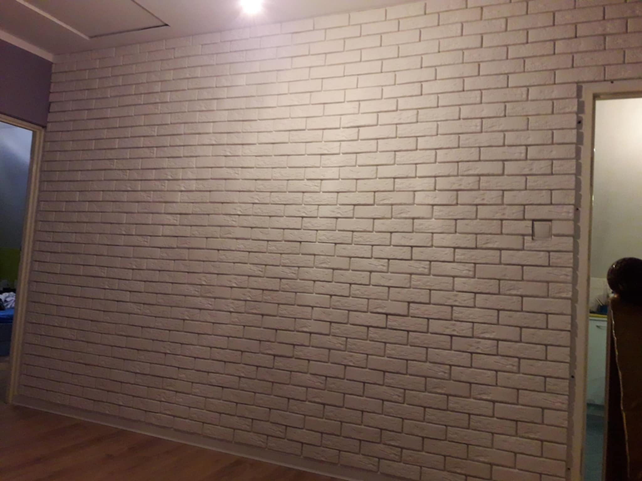 Ściana wykończona dekoracyjną, białą cegłą w pomieszczeniu z drewnianą podłogą. Widoczne drzwi po prawej stronie i fragment sufitu z klapą rewizyjną.