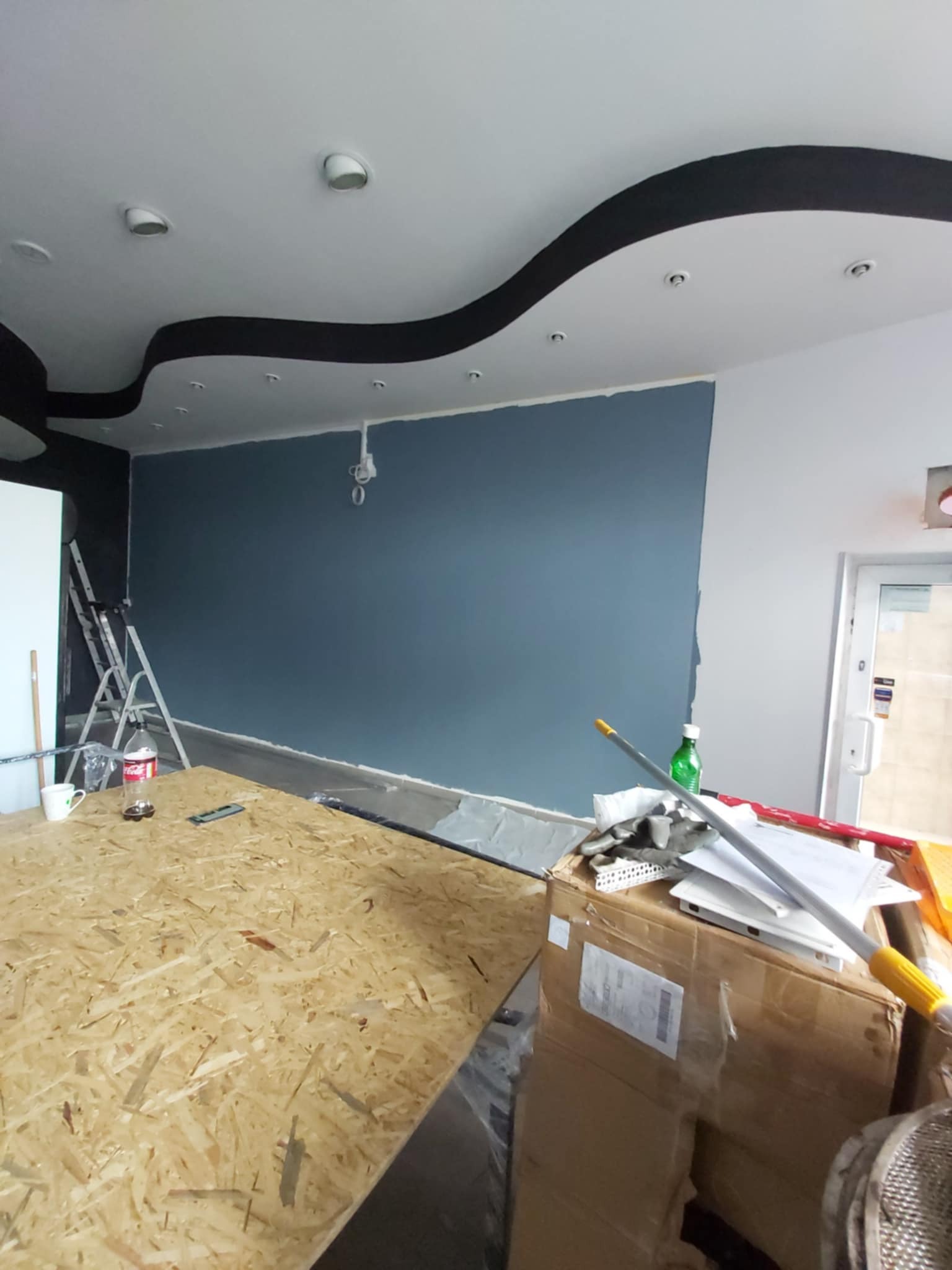 Niebieska ściana w trakcie malowania, widoczny fragment sufitu z dekoracyjnym wykończeniem, narzędzia malarskie i drabina. Częściowo zakryte podłoże robocze.