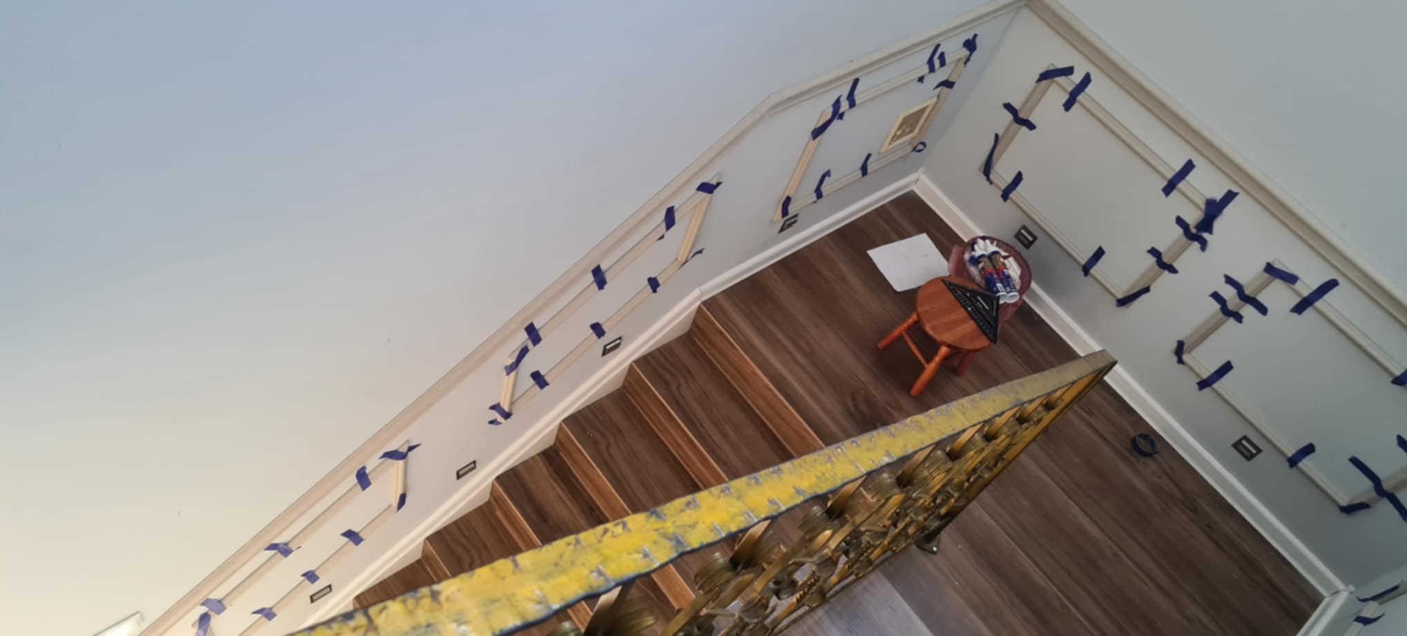 Montaż listew dekoracyjnych na ścianie klatki schodowej, przyklejonych niebieską taśmą malarską. Widok z góry na drewniane schody i fragment metalowej balustrady.
