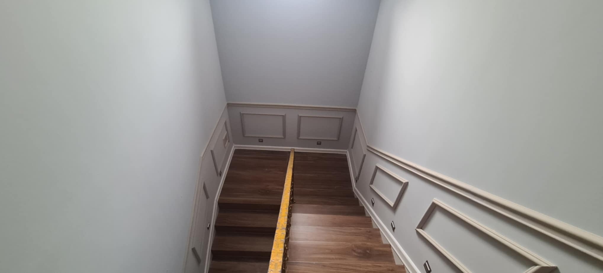 Schody z drewnopodobnymi panelami, szare ściany z dekoracyjnymi listwami i oświetleniem punktowym. Widok z góry, z żółtą barierką zabezpieczającą.